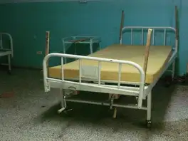Cama de hospital con un colchón de espuma sin forro y tablas de madera amarradas a las barandas.