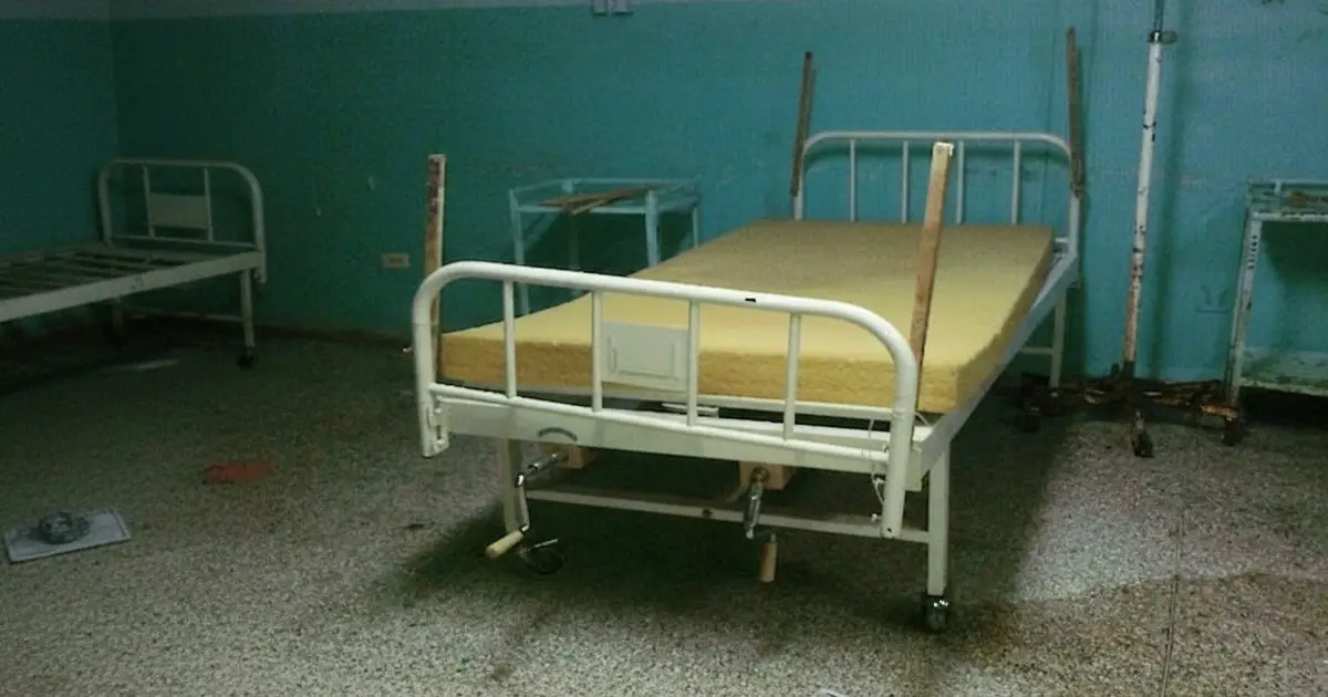 Cama de hospital con un colchón de espuma sin forro y tablas de madera amarradas a las barandas.