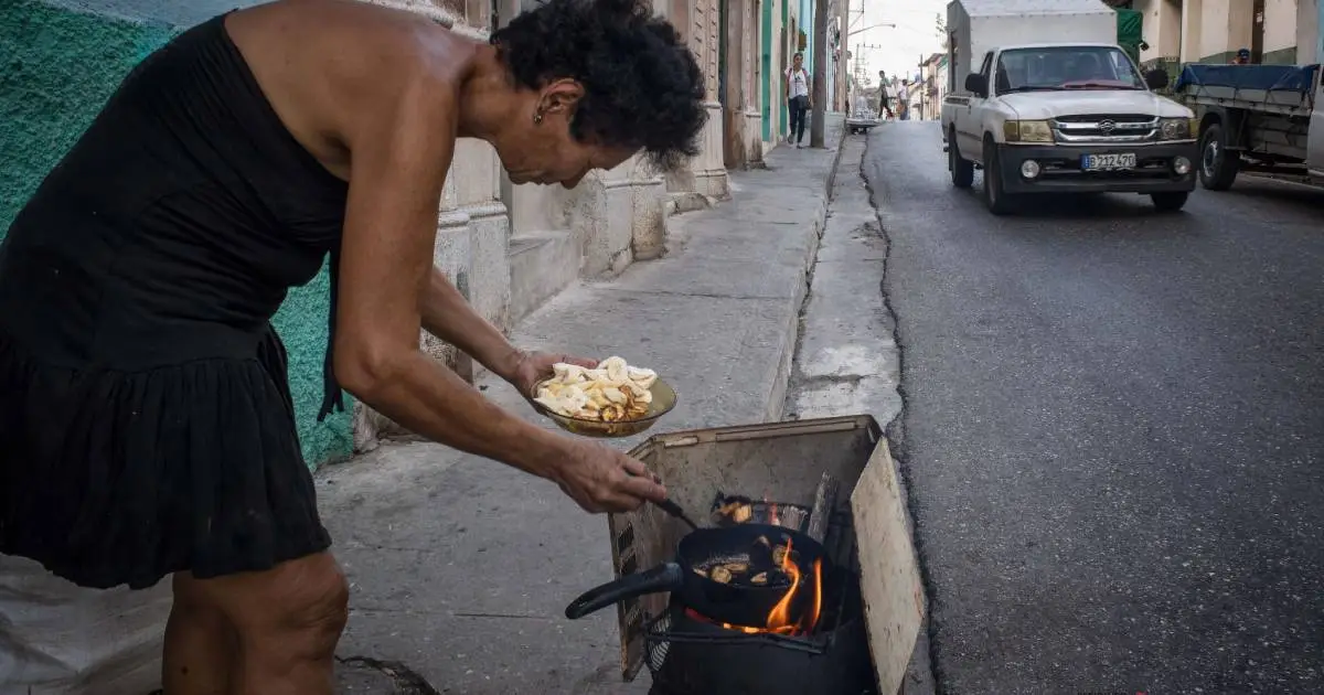 Una mujer fríe rodajas de plátano en una sartén sobre un fogón de leña improvisado en la acera de una calle.