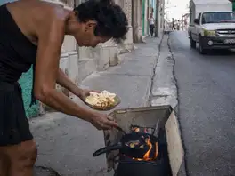 Una mujer fríe rodajas de plátano en una sartén sobre un fogón de leña improvisado en la acera de una calle.